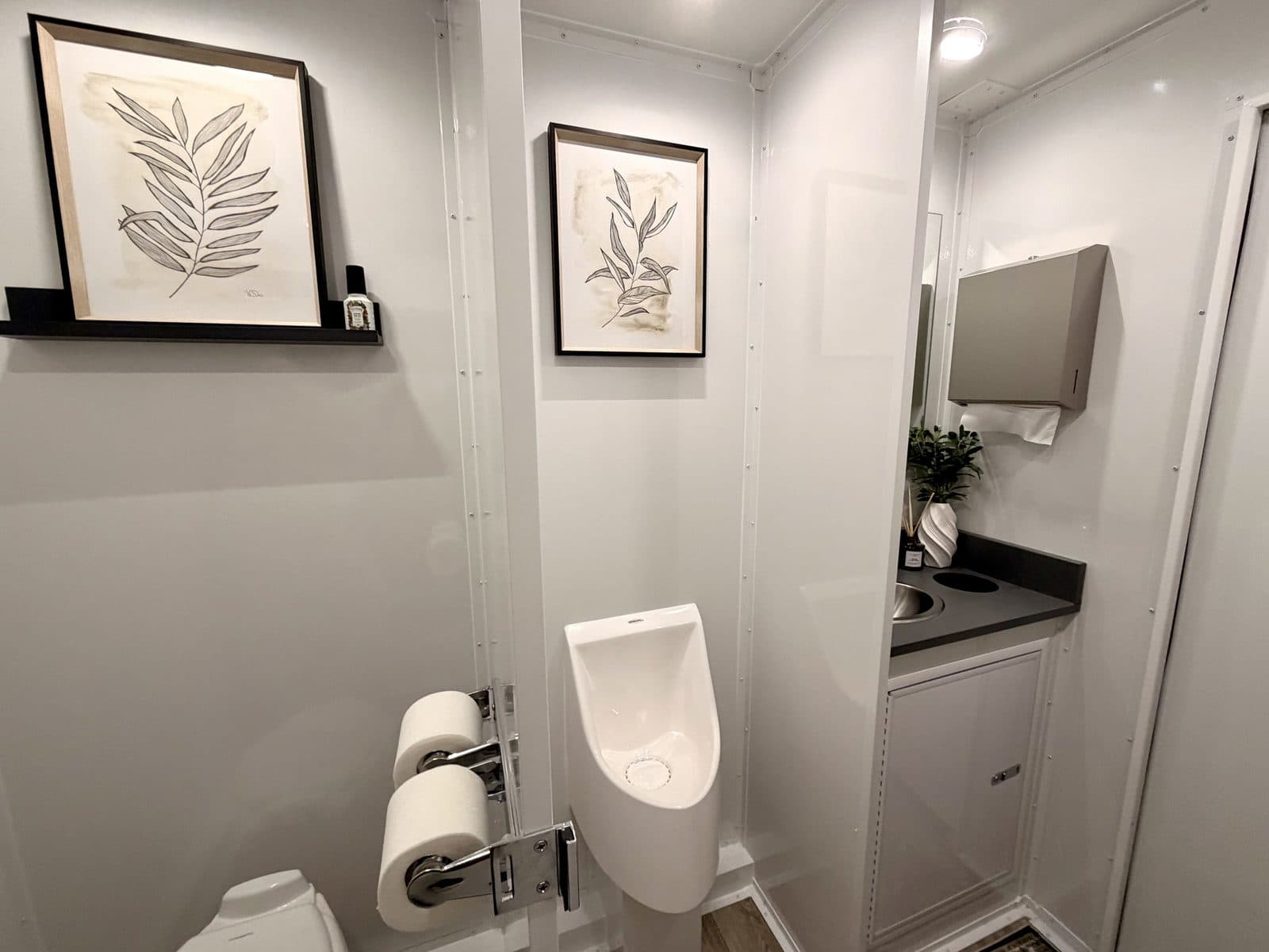 Men's station inside a luxury restroom trailer with urinal, framed art, stocked toilet paper, and a stainless sink