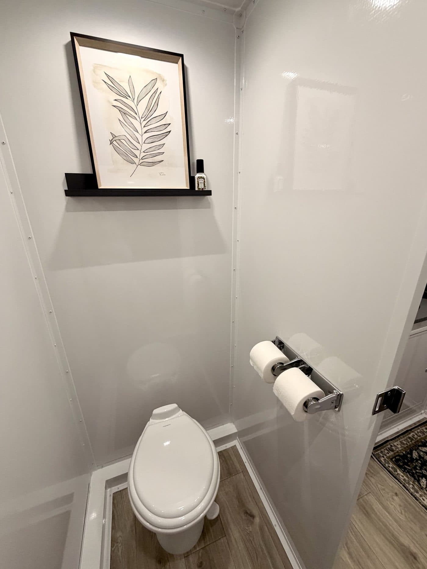 Private stall inside a luxury restroom trailer with a flushing toilet, framed botanical art, and wood-look flooring